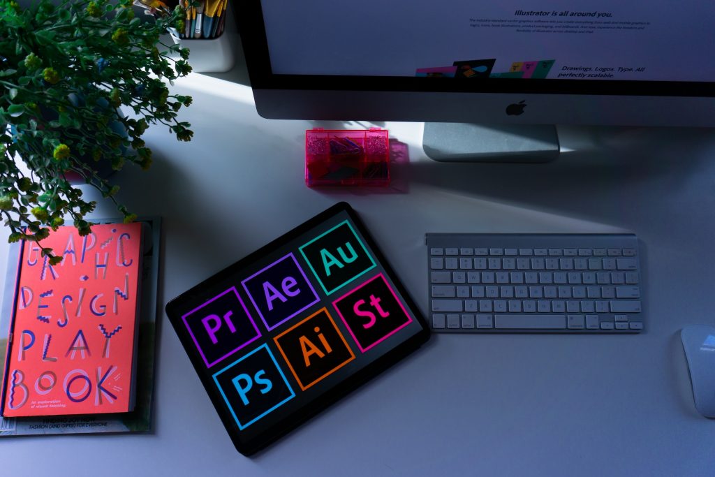 image of computer with a tablet of several Adobe Creative Cloud Suite programs on a table next to a book of design